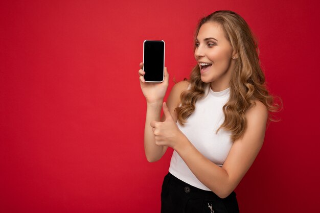 Bella giovane donna bionda che indossa t-shirt bianca isolata su sfondo rosso con spazio copia che tiene smartphone che mostra telefono in mano con schermo vuoto per mockup