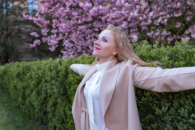 Bella giovane donna bionda con i capelli ricci nel giardino in fiore di ciliegio che guarda in alto e gode del giorno di primavera ragazza all'aperto