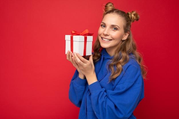 Bella giovane donna bionda felice isolata sopra il muro di sfondo colorato che indossa abiti casual alla moda che tengono il contenitore di regalo e che guarda l'obbiettivo.