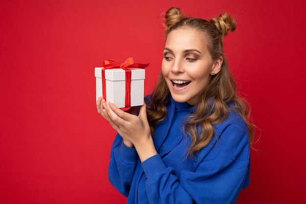 Bella giovane donna bionda felice isolata sopra la parete variopinta del fondo che indossa vestiti casuali alla moda che tengono il contenitore di regalo e che guarda al lato.