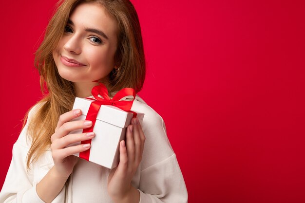 Bella giovane donna bionda felice isolata sopra la parete variopinta del fondo che indossa vestiti casuali alla moda che tengono scatola regalo e che guarda l'obbiettivo.