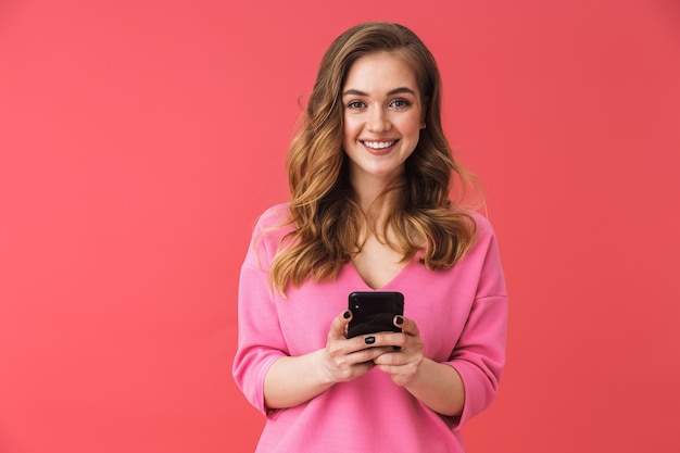 Bella giovane donna bionda in piedi isolata sul muro rosa, usando il telefono cellulare