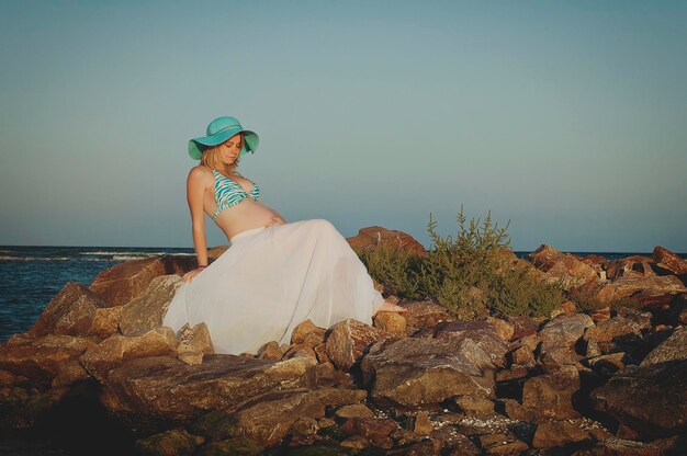 Bella giovane donna bionda incinta elegante con un cappello blu cammina sulla spiaggia, mare al tramonto.