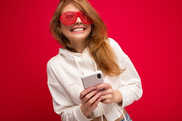 Bella giovane donna bionda sorridente che indossa felpa con cappuccio bianca alla moda e occhiali colorati divertenti