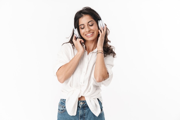 Bella giovane donna bruna allegra che indossa abiti casual in piedi isolato su un muro bianco, ascoltando musica con cuffie wireless