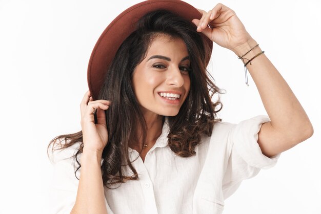 Bella giovane donna bruna allegra che indossa abiti casual in piedi isolato su un muro bianco, posando in un cappello