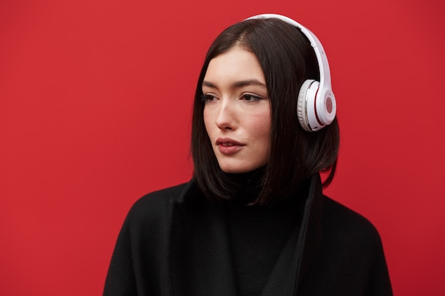 Bella giovane donna bruna che indossa cappotto autunnale in piedi isolata sul rosa, ascoltando musica con cuffie wireless