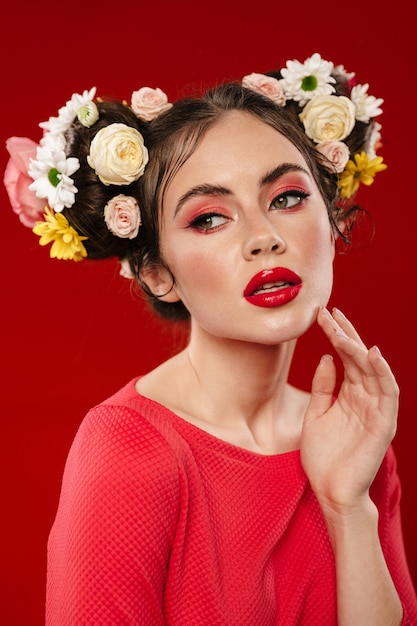 Bella giovane donna bruna con un'incredibile acconciatura floreale che indossa il trucco in posa isolata sul muro rosso