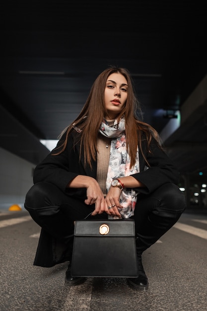 Bella giovane donna bruna in abiti eleganti di eleganza nera con borsa in pelle alla moda è seduta sull'asfalto in città. Ragazza europea moderna alla moda in posa sulla strada in strada.