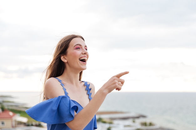 Bella giovane donna bruna sorridente in piedi sulla terrazza bianca e puntare il dito contro qualcosa