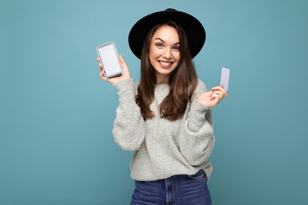 Bella giovane donna bruna sorridente positiva che indossa cappello nero e maglione grigio isolato sopra