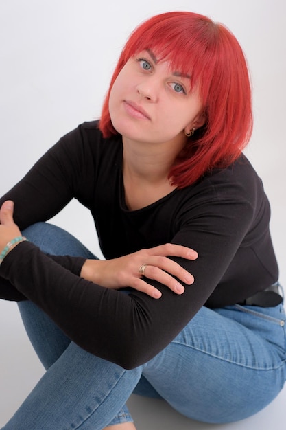 Bella giovane donna carina con l'acconciatura arancione in jeans seduta in posa in studio su sfondo bianco