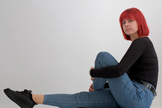 Bella giovane donna carina con l'acconciatura arancione in jeans seduta in posa in studio su sfondo bianco