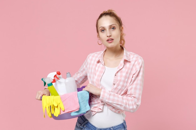 Bella giovane donna casalinga in camicia a scacchi bacinella con bottiglie di detersivo che lavano i detergenti mentre fanno i lavori domestici isolati su sfondo rosa studio. Concetto di pulizia. Guardando la fotocamera.