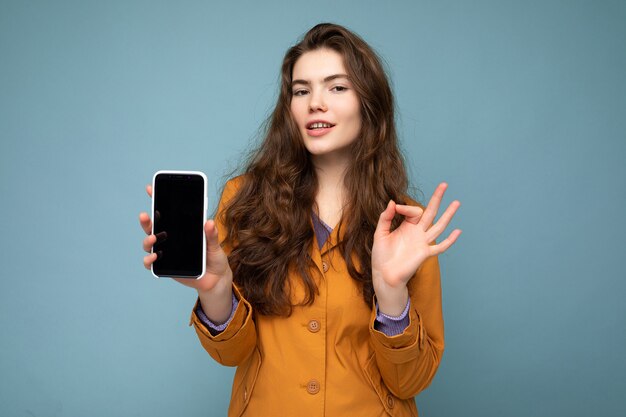 Bella giovane donna castana che indossa giacca arancione isolato su sfondo blu tenendo in mano e mostrando mobile