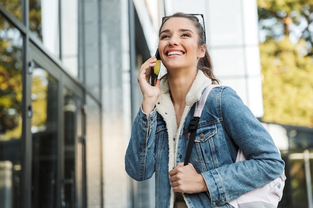 Bella giovane donna castana che indossa una giacca, portando zaino camminando all'aperto, parlando al telefono cellulare