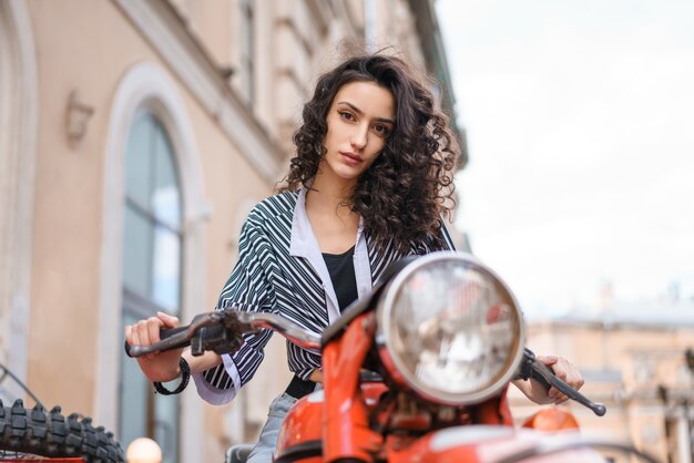 Bella giovane donna castana con capelli ricci che si siede su una motocicletta rossa