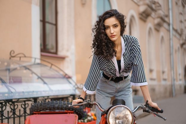 Bella giovane donna castana con capelli ricci che si siede su una motocicletta rossa