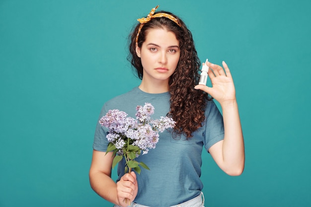 Bella giovane donna castana con il mazzo di lillà fiorito che mostra lo spray antiallergico per il naso che usa per curare l'allergia ai pollini