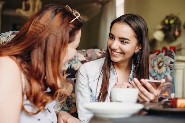 Bella giovane donna caucasica con capelli lunghi scuri guardando la sua amica sorridente mentre si tiene uno smartphone mentre è seduto nel ristorante.