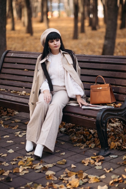 Bella giovane donna caucasica con lunghi capelli scuri in camicetta bianca, pantaloni grigi, cappello bianco e cappotto grigio con borsa marrone nella foresta