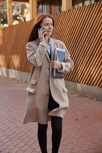 Bella giovane donna caucasica dalla testa rossa in cappotto beige con i capelli lunghi che tiene il laptop all'aperto, chiamando tramite smartphone. attraente ritratto femminile in autunno. stile di vita autunnale, lavoro freelance, moda