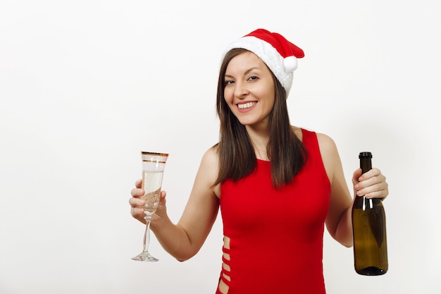 Bella giovane donna caucasica felice con un sorriso affascinante in abito rosso e cappello di Natale in piedi con bicchiere e bottiglia di champagne su sfondo bianco. Ragazza di Santa isolata. Concetto di vacanza di Capodanno.