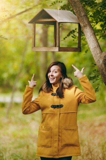 Bella giovane donna caucasica felice sorridente dai capelli castani in cappotto giallo, jeans, stivali nella foresta verde. Modello di moda con foglie autunnali in piedi sotto mangiatoie per uccelli nel parco di inizio autunno all'aperto.