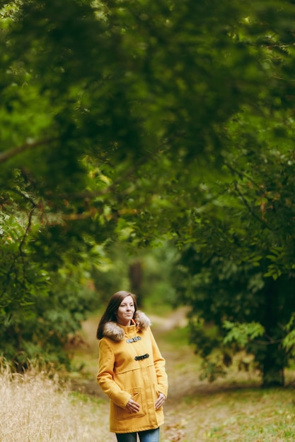 Bella giovane donna caucasica felice sorridente dai capelli castani in cappotto giallo, jeans, stivali nella foresta verde. Modello femminile di moda con foglie d'oro autunnali in piedi e camminando nel parco all'inizio dell'autunno all'aperto.