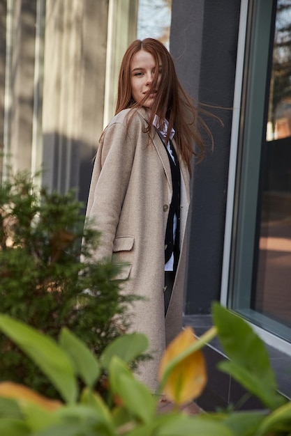 Bella giovane donna caucasica in cappotto beige con capelli lunghi in piedi all'aperto dietro piante verdi
