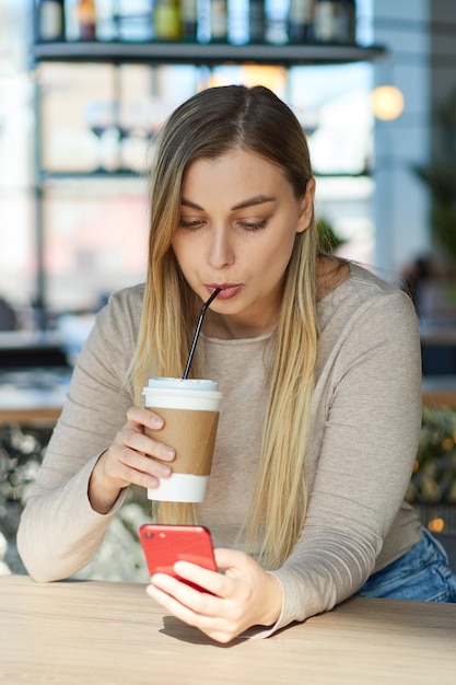 Bella giovane donna che beve caffè in un caffè da una tazza di carta utilizzando un telefono cellulare