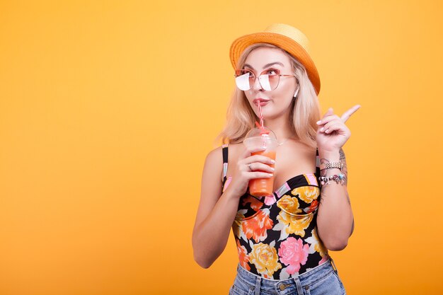 Bella giovane donna che beve succo d'arancia fresco con occhiali da sole e cappello in studio su sfondo giallo