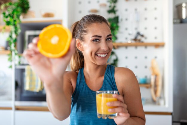 Bella giovane donna che beve succo d'arancia fresco in cucina dieta sana giovane donna felice con un bicchiere di succo e arancia al tavolo in cucina