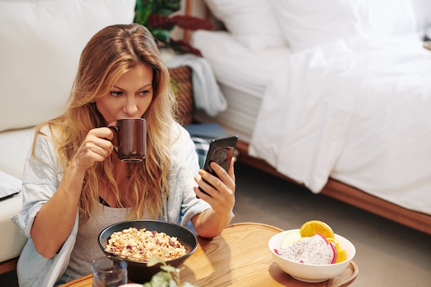 Bella giovane donna che controlla i social media sullo smartphone quando mangia la sua colazione sana e beve caffè a casa