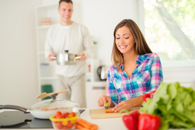 Bella giovane donna che cucina un pasto sano nella cucina domestica. Il suo ragazzo dietro, con in mano la pentola. Messa a fuoco selettiva. Concentrati sul primo piano.