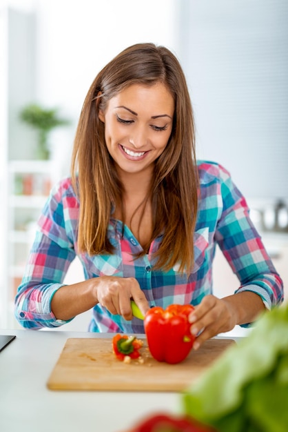 Bella giovane donna che fa pasto sano nella cucina domestica. Sta tagliando il peperoncino sul tagliere della cucina.