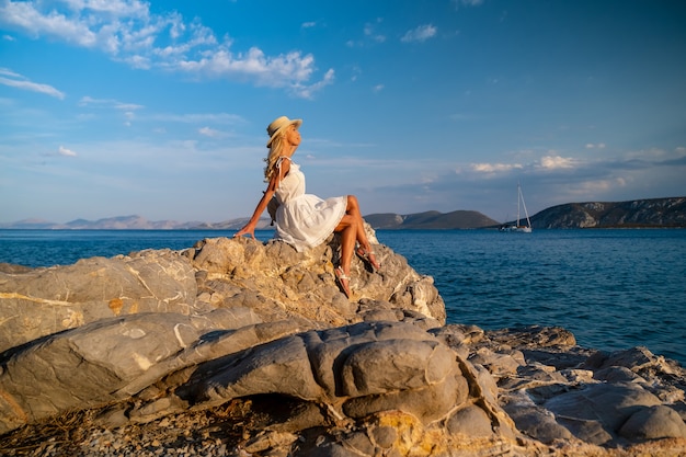 Bella giovane donna che indossa cappello da traino seduto tra le rocce in abito bianco in riva al mare