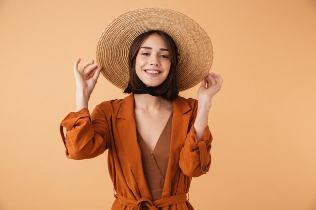 Bella giovane donna che indossa cappello di paglia e vestito estivo in piedi isolato sul muro beige, in posa