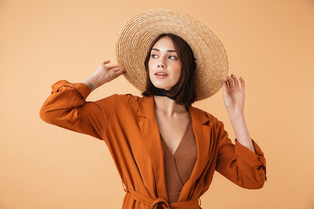 Bella giovane donna che indossa cappello di paglia e vestito estivo in piedi isolato sul muro beige, in posa