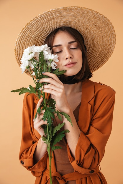 Bella giovane donna che indossa un cappello di paglia e un vestito estivo in piedi isolato su un muro beige, con in mano un bouquet di camomilla