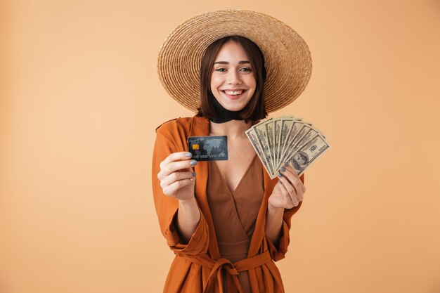 Bella giovane donna che indossa un cappello di paglia e un vestito estivo in piedi isolato su un muro beige, con in mano una carta di credito palstica, che mostra banconote in denaro