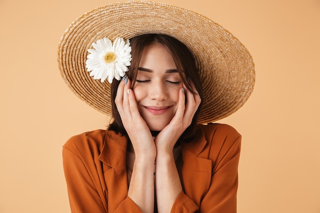 Bella giovane donna che indossa un cappello di paglia e un vestito estivo in piedi isolato su un muro beige, in posa con fiori di camomilla