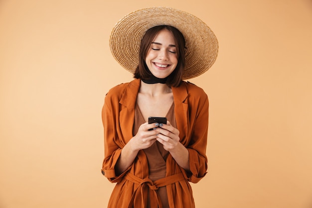 Bella giovane donna che indossa un cappello di paglia e un vestito estivo in piedi isolato su un muro beige, usando il telefono cellulare