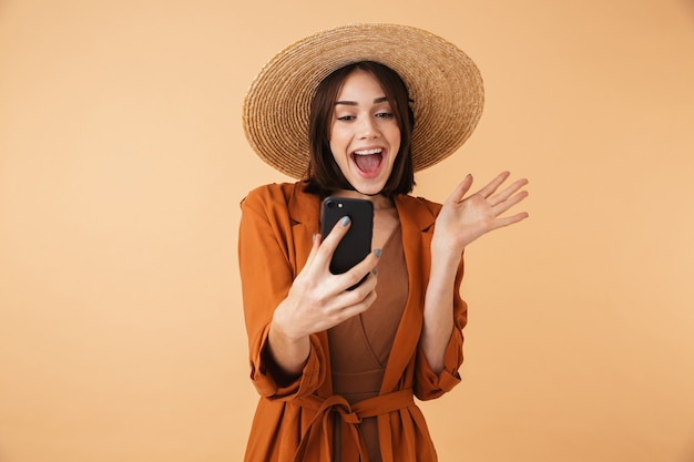 Bella giovane donna che indossa un cappello di paglia e un vestito estivo in piedi isolato sul muro beige, facendo un selfie