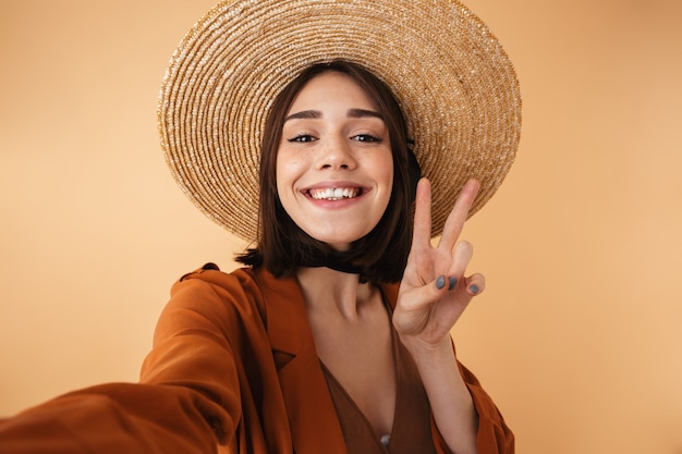 Bella giovane donna che indossa un cappello di paglia e un vestito estivo in piedi isolato sul muro beige, facendo un selfie