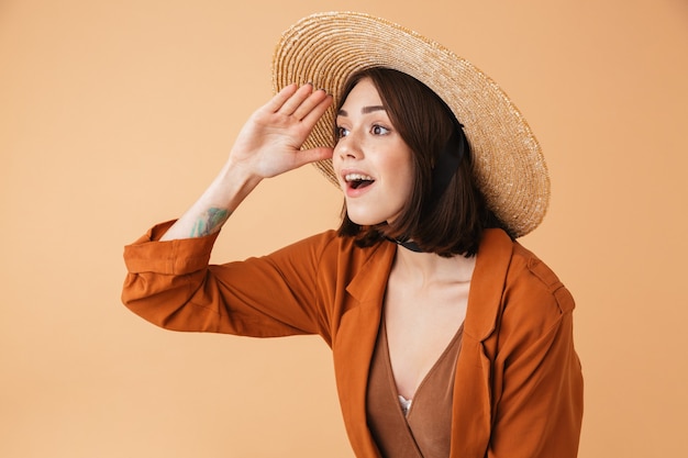 Bella giovane donna che indossa un cappello di paglia e un vestito estivo in piedi isolato sul muro beige, gridando forte