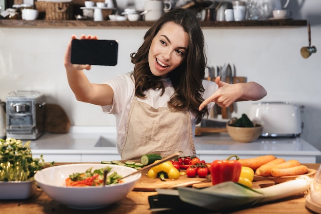 Bella giovane donna che indossa un grembiule che cucina una sana insalata in cucina a casa, facendo un selfie con il cellulare