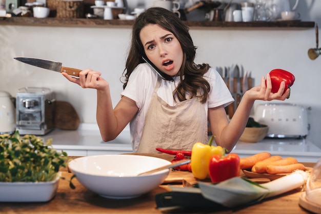 Bella giovane donna che indossa un grembiule che cucina una sana insalata in cucina a casa, utilizzando il telefono cellulare durante la cottura