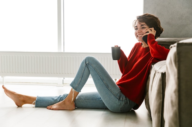 Bella giovane donna che indossa un maglione seduto su un pavimento a casa, bere il tè, appoggiato su un divano