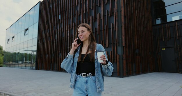 Bella giovane donna che indossa una giacca di jeans e utilizza uno smartphone moderno mentre si cammina in pausa in città Donna che parla al telefono con gli amici gioendo sorridendo ridendo e bevendo caffè